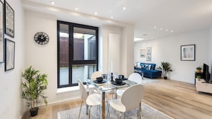 A modern living room with a glass dining table, white chairs, a blue sofa, and a TV.