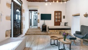 Une pièce chaleureuse avec un mur en pierre, une porte en bois, un lit avec une tête de lit, un fauteuil gris, une petite table avec un vase et un téléviseur fixé au mur.