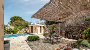 Un espace aménagé au bord de la piscine, doté d’un sol en pierre, d’un coin salon couvert avec des chaises et une table, ainsi que d’un mur en pierre.