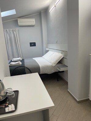 Een compacte hotelkamer met een bed, nachtkastje en een klein bureau met een dienblad met waterkoker, kopjes en theezakjes.