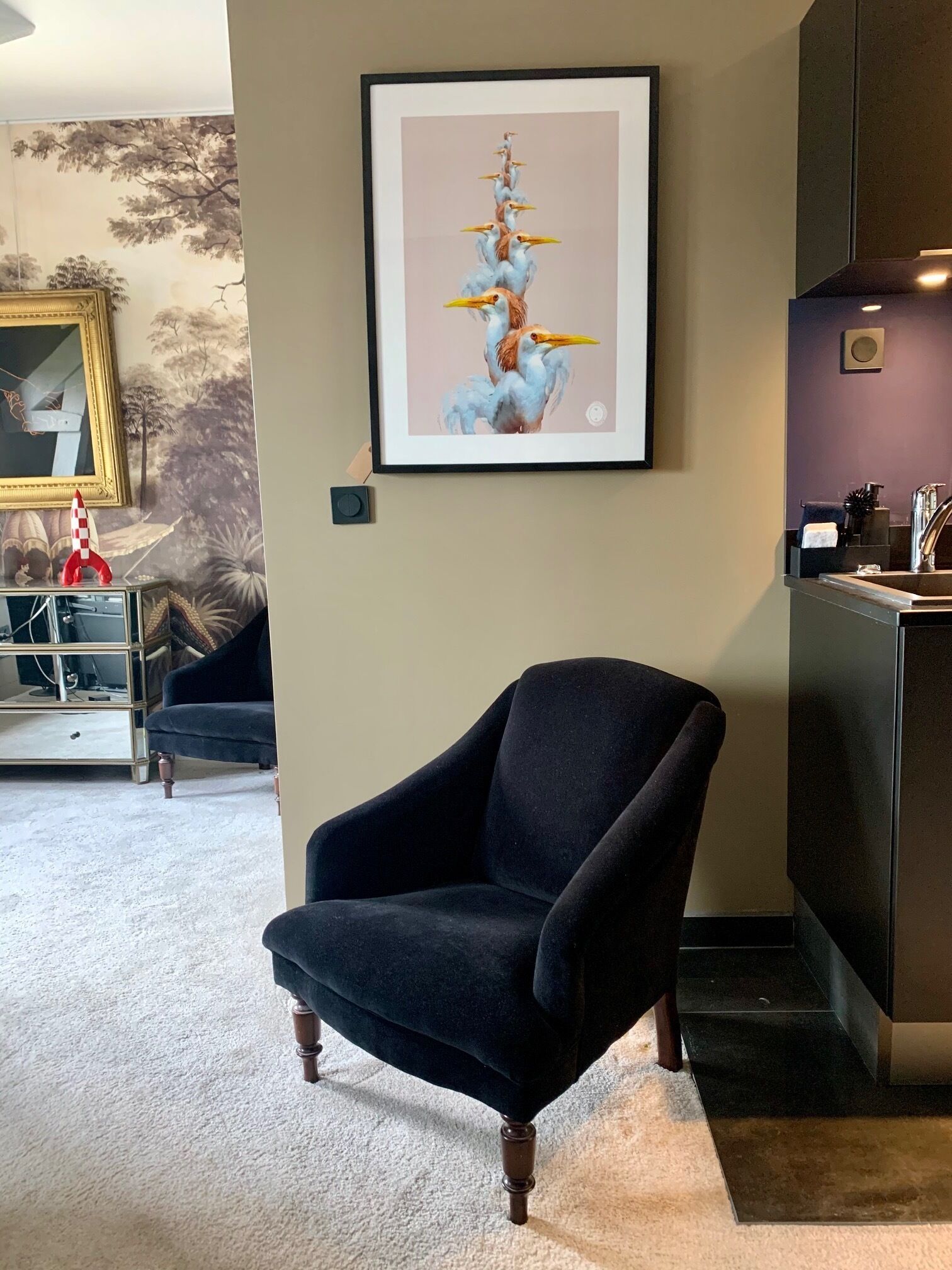 A room with a framed artwork of birds, a dark armchair, a small table with a red and white object, and a mirror on the wall.