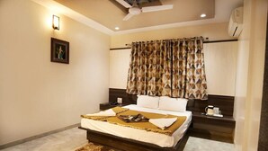 A bedroom with a bed, bedside tables, a nightstand, a ceiling fan, a wall-mounted air conditioner, and a patterned curtain. - Hotel Sai Suraj Park (Rahata)