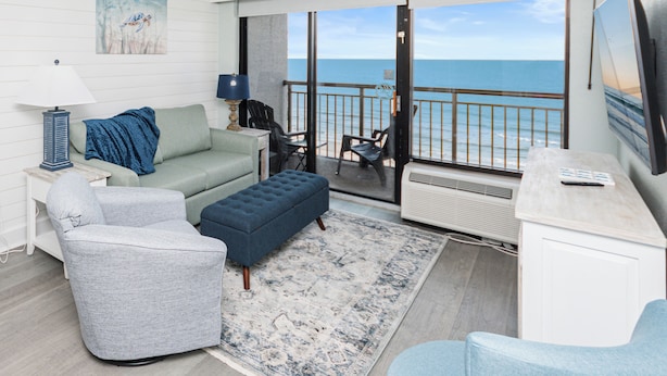 Een moderne woonkamer met een bank, fauteuil, voetenbankje en uitzicht op de oceaan door grote ramen.