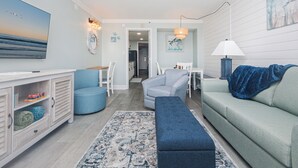 Een woonkamer met een lichtblauwe bank, een blauwe fauteuil, een witte eettafel en een blauwe bank. Aan de muur hangt een groot schilderij en op de tafel staat een lamp.