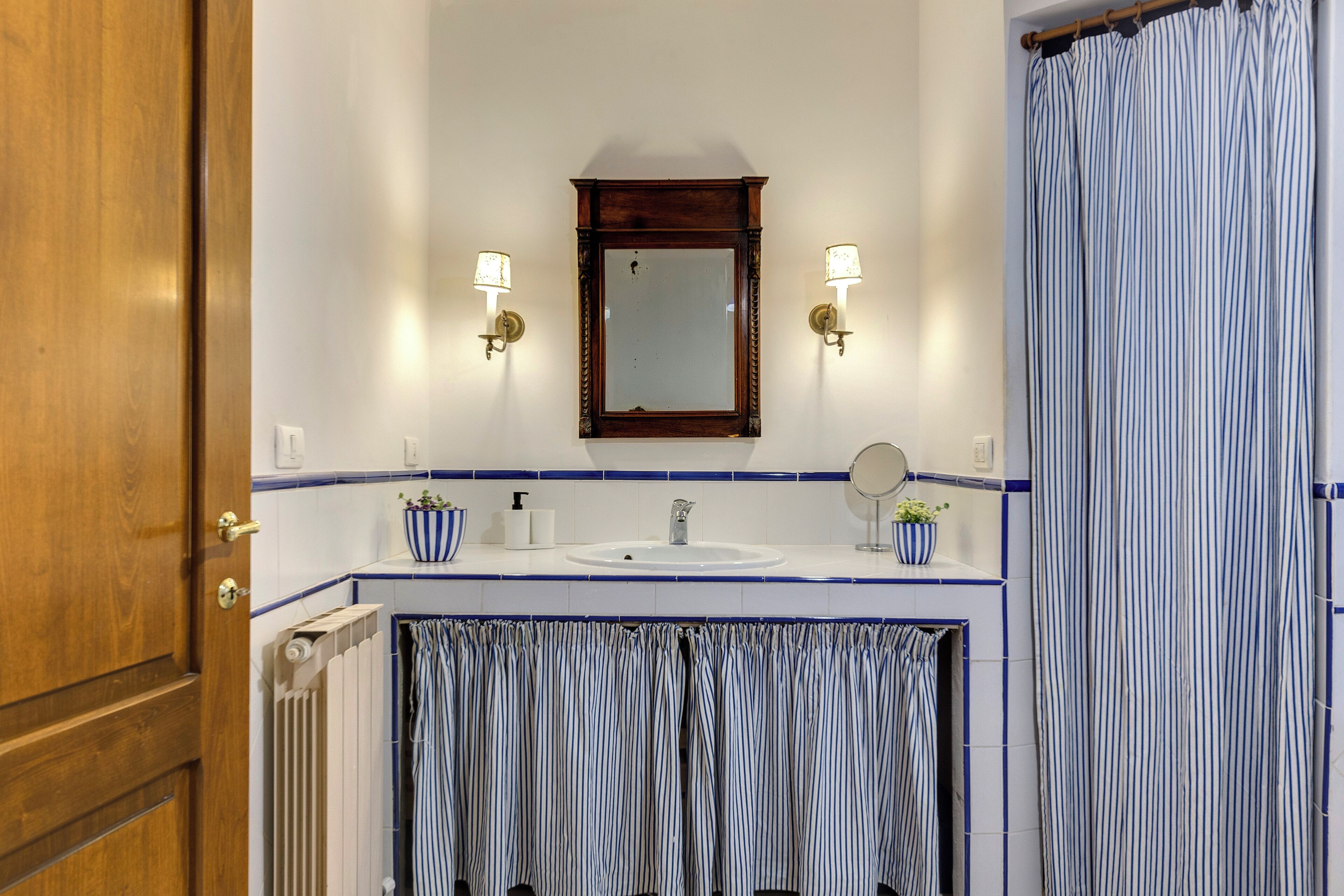 Un bagno con porta in legno, pareti piastrellate di bianco, un lavandino bianco, uno specchio, due luci a parete e una tenda da doccia a righe.