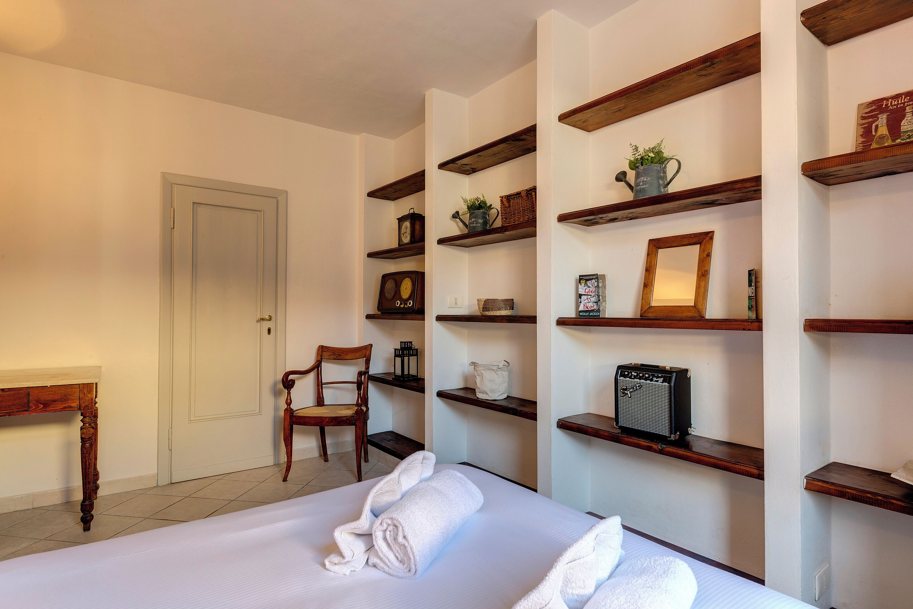 Una habitación de hotel con una cama bien tendida, estantes de madera con varios objetos, una silla y una puerta.