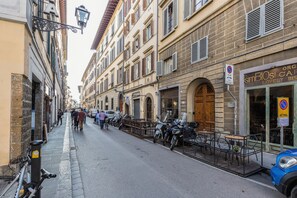 Una strada con scooter e biciclette parcheggiate, un caffè con tavolini all'aperto e un edificio con un'insegna che riporta la scritta "SIMBIOS".