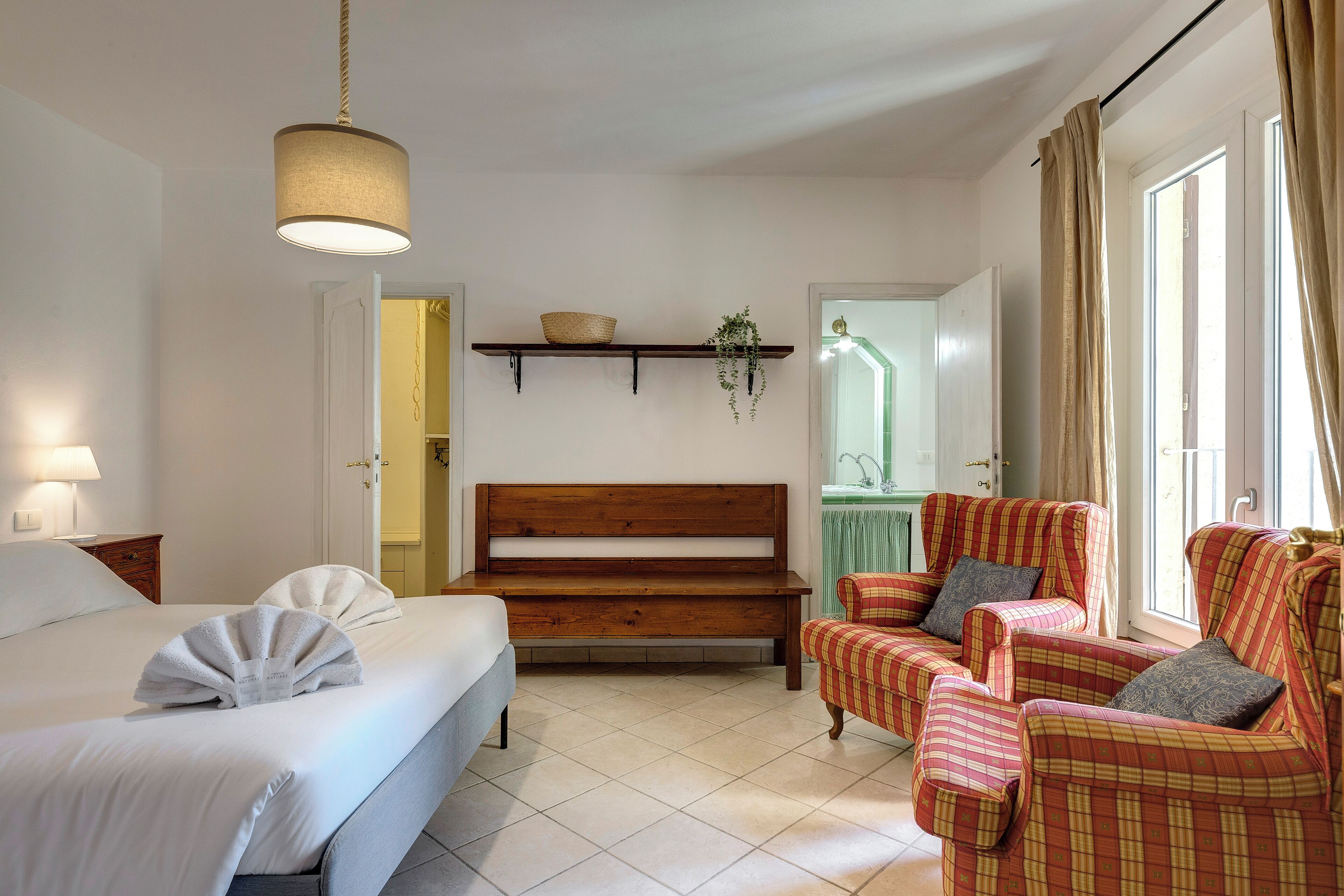 A bedroom with a bed, a bench, two armchairs, a hanging plant, and a shelf with a basket.