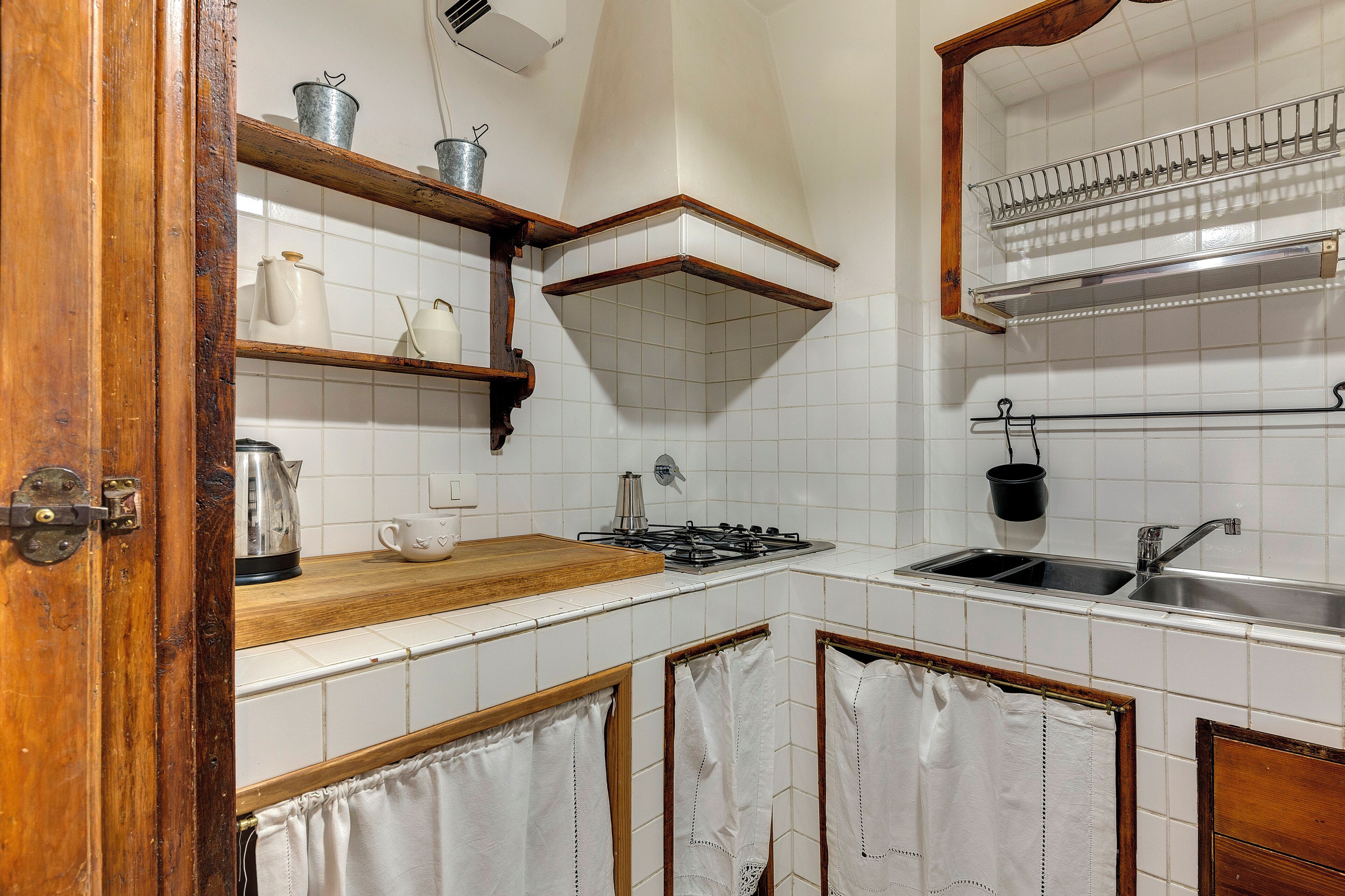 Una cocina con azulejos blancos, estantes de madera, una estufa a gas y un lavamanos.
