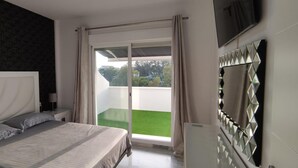 A bedroom with a large bed, a nightstand, a TV, and a sliding glass door leading to a balcony with a green lawn. - Marbella Oasis Villa With 6 Bedrooms (Marbella)