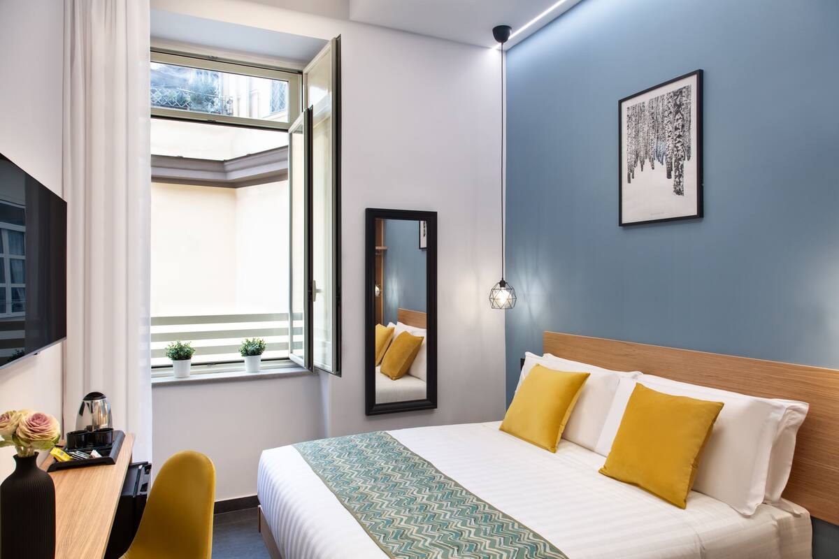 A hotel room with a bed, a desk, a chair, a window, a mirror, and a wall-mounted picture.