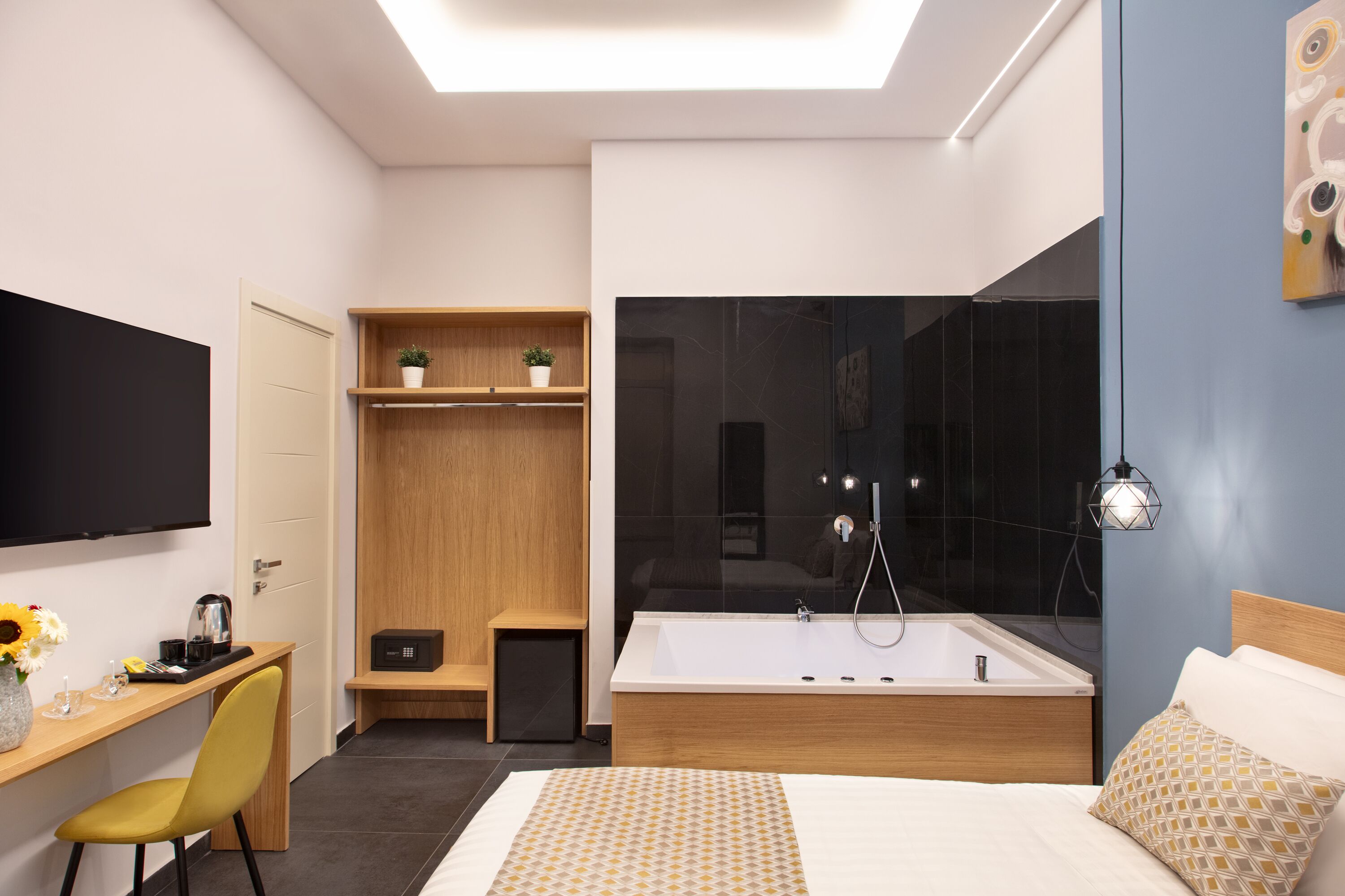 A modern hotel room with a large bed, a desk with a chair, a shower area, and a TV mounted on the wall.
