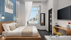 A modern hotel room with a large bed, a wooden bedside table, a built-in TV, a bathroom with a bathtub, and a view of a cityscape through a large window. - MAIN - Duomo (Naples)