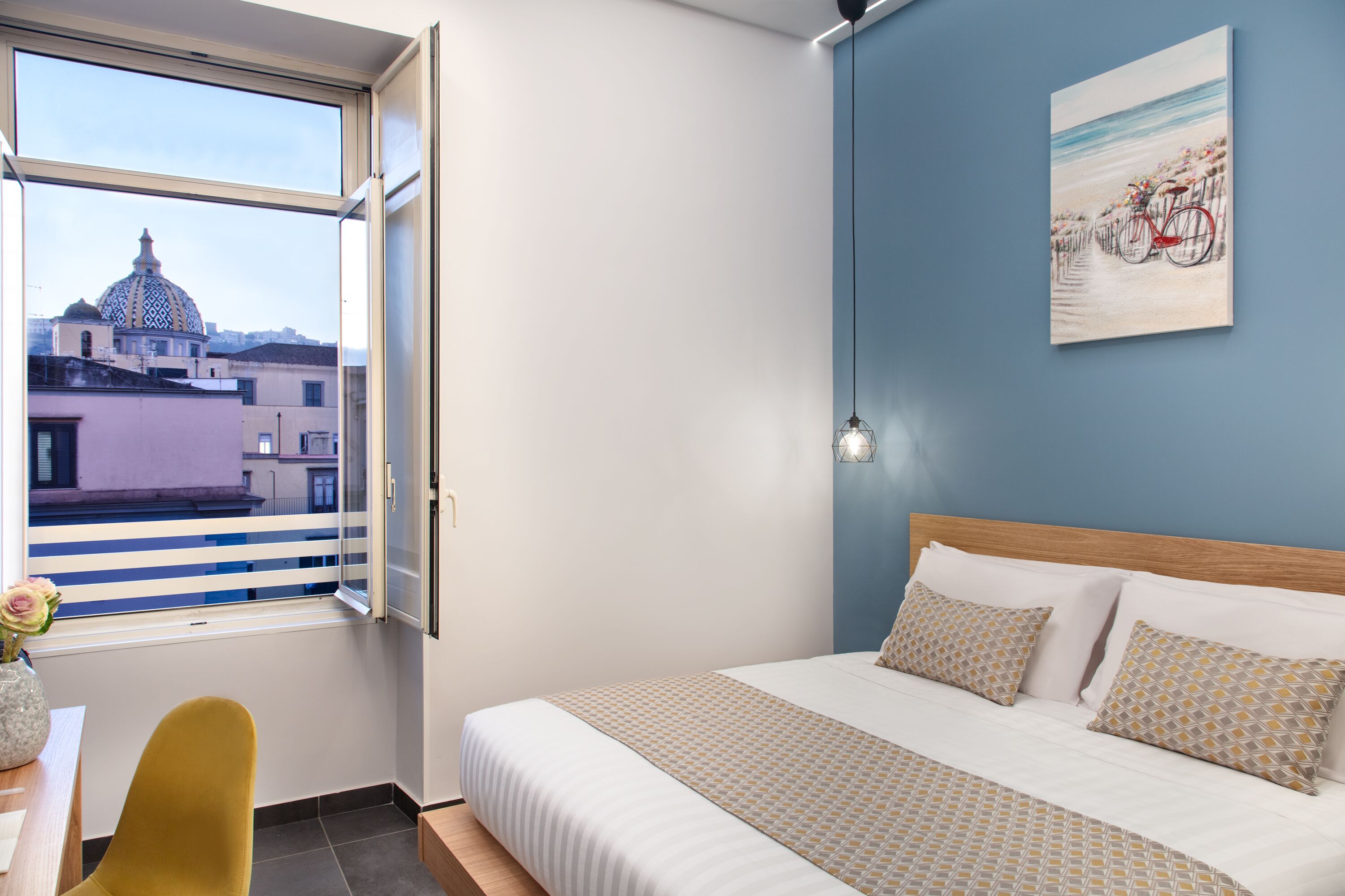 A hotel room with a bed, a window with a view of a building, a painting on the wall, and a vase with flowers on the table.