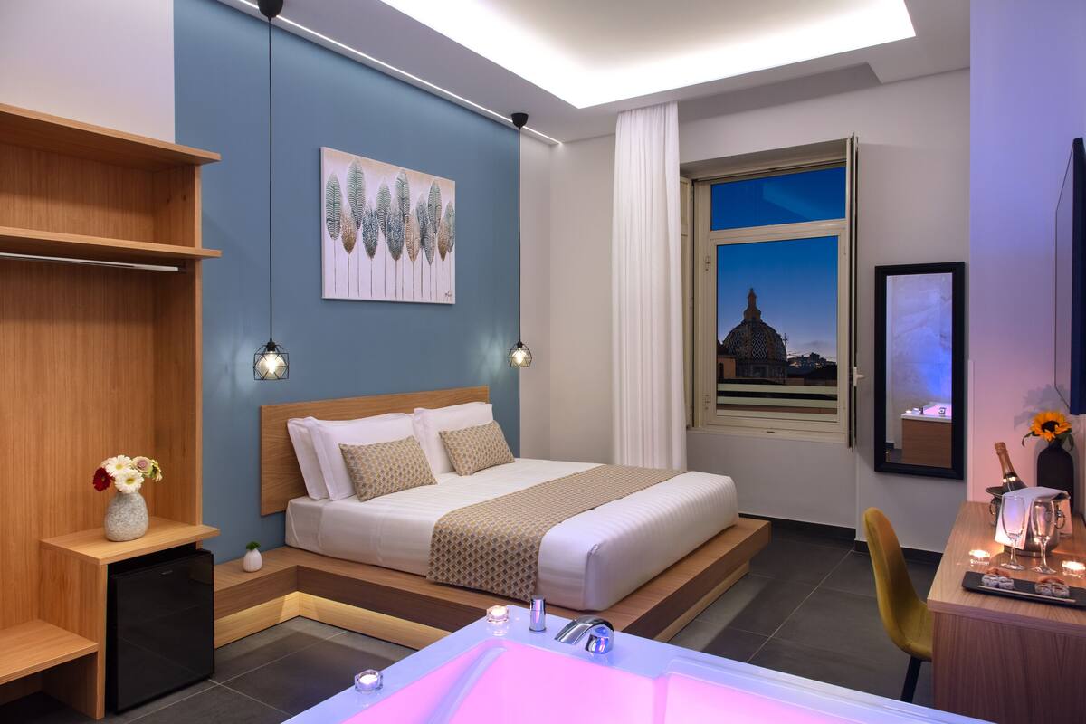 A modern hotel room with a large bed, bedside tables, a desk, a bathroom with a bathtub, and a cityscape view.