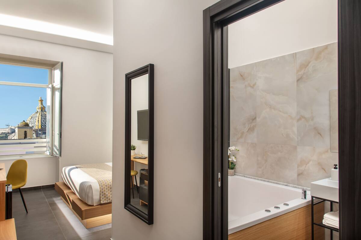 A modern hotel room with a large bed, a desk, a chair, a bathroom with a large mirror, and a view of a cityscape through a window.