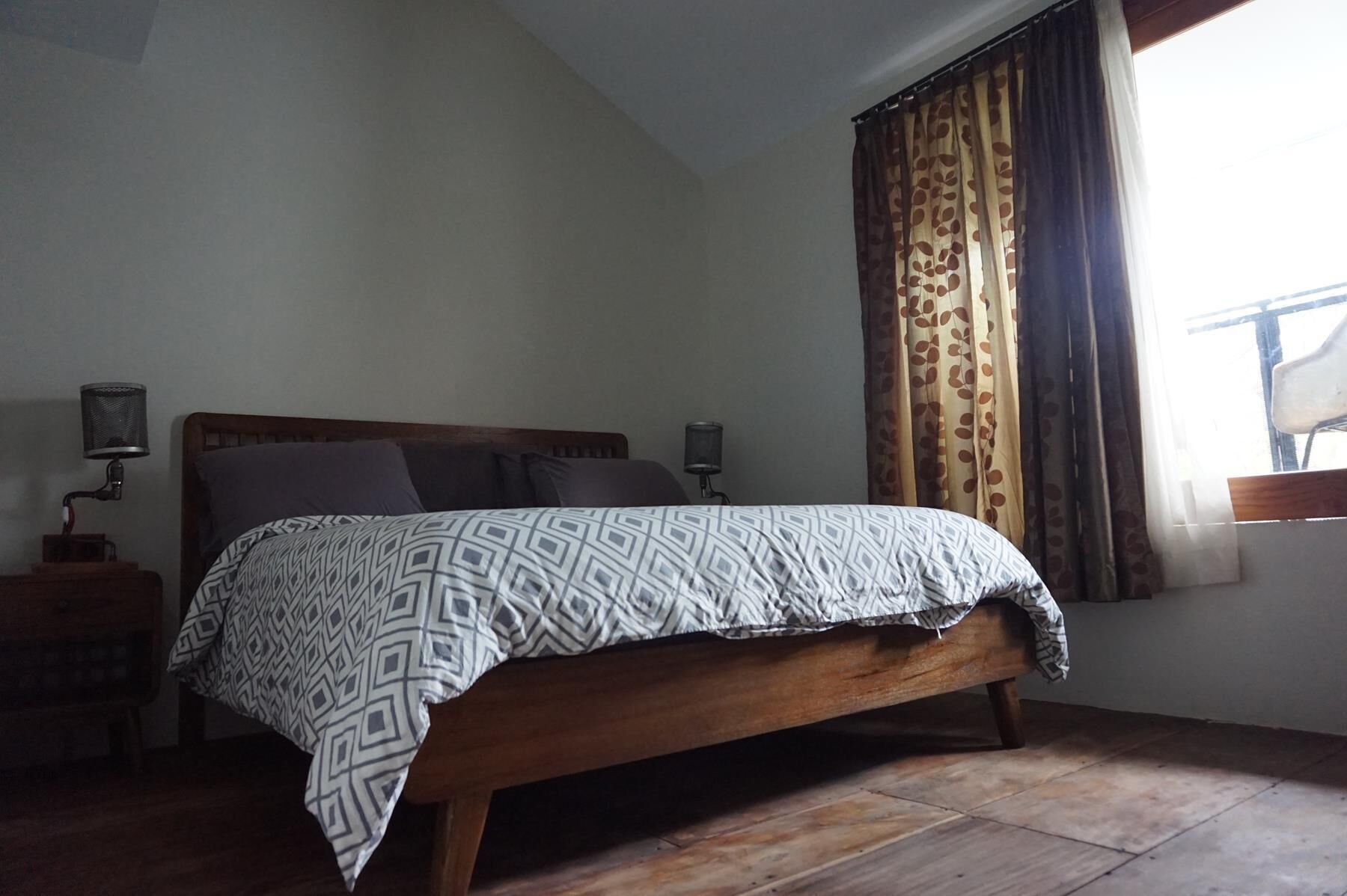Ein Schlafzimmer mit einem hölzernen Bett, gemusterten Betttextilien, zwei Nachttischlampen und einem Fenster mit Vorhängen.