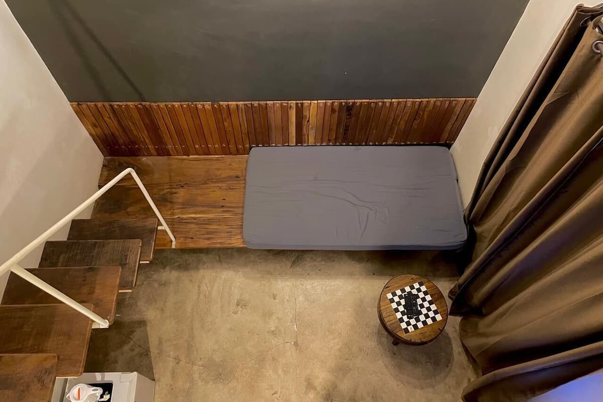 A small room with a wooden floor, a grey mattress, a round wooden table with a checkered top, and a staircase with wooden steps and white railings.