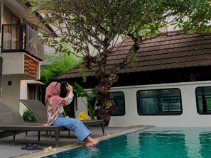 Une personne se détend au bord d’une piscine, avec un arbre et un bâtiment en arrière-plan.