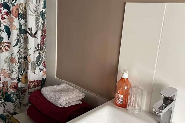 A bathroom with a floral shower curtain, a white sink, a bottle of lavender body wash, a glass, and a faucet. - The Dairy - Seaside Couples Escape in Whitstable (Whitstable)