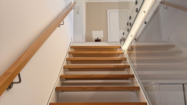 Eine moderne Treppe mit Holzboden und Glasgeländern, die zu einer Zwischenebene mit einer Tür und einem kleinen Tisch führt.