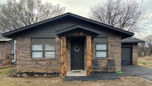 A single-story brick house with a dark roof, a front door with a wreath, and two windows with shutters.