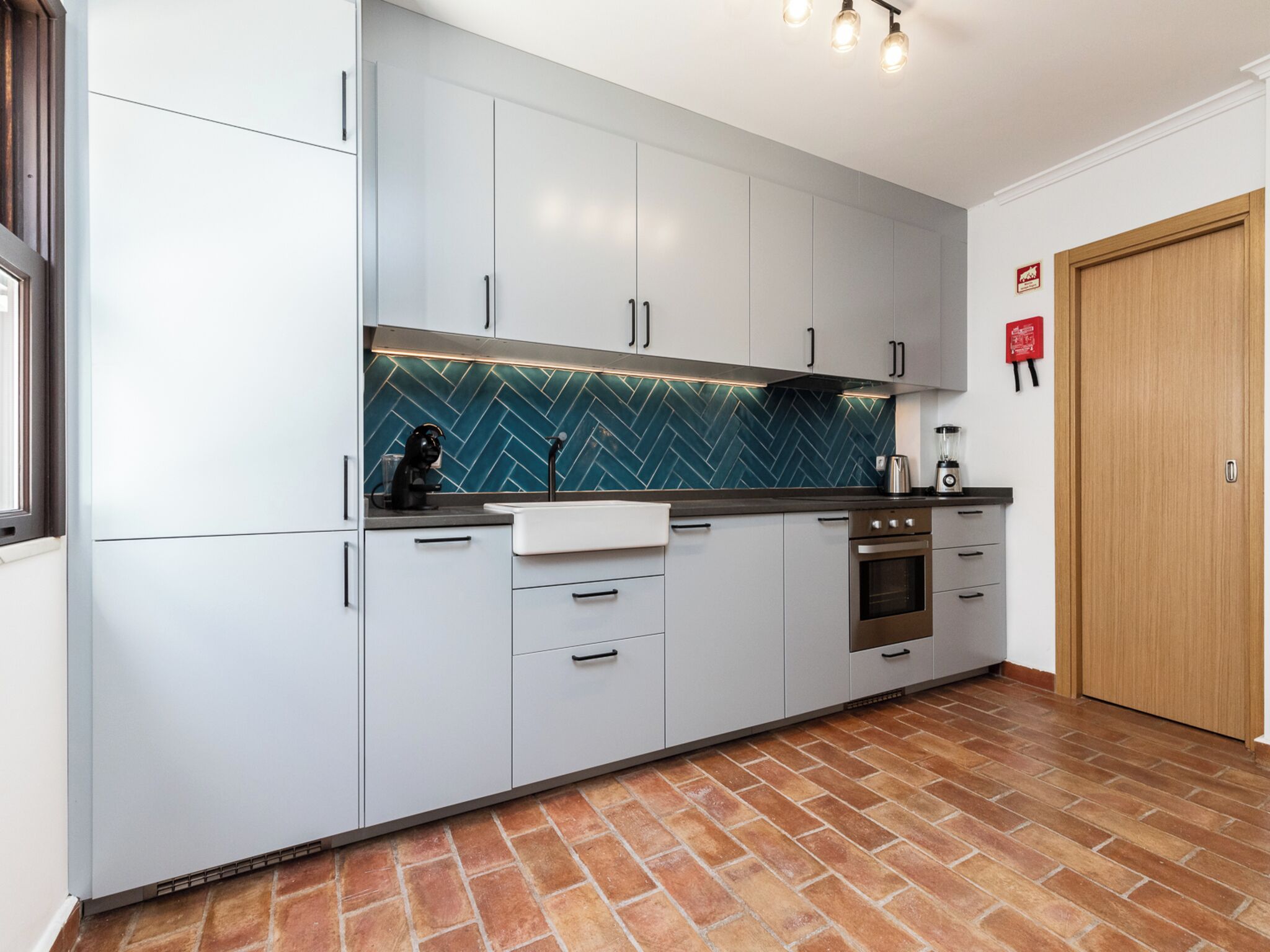 Una cucina moderna con mobili bianchi, una parete di fondo a spina di pesce e un pavimento in mattoni.
