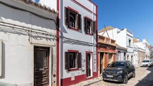 A street with a two-story building with a red and white facade, a parked car, and a cobblestone road. - Zinnia Villa, Tavira, Algarve (Tavira)