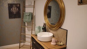 A bathroom with a wooden vanity, a round mirror, a white sink, a towel rack, and a decorative wall piece. - Au petit bonheur de Séraphine (saint aubin)