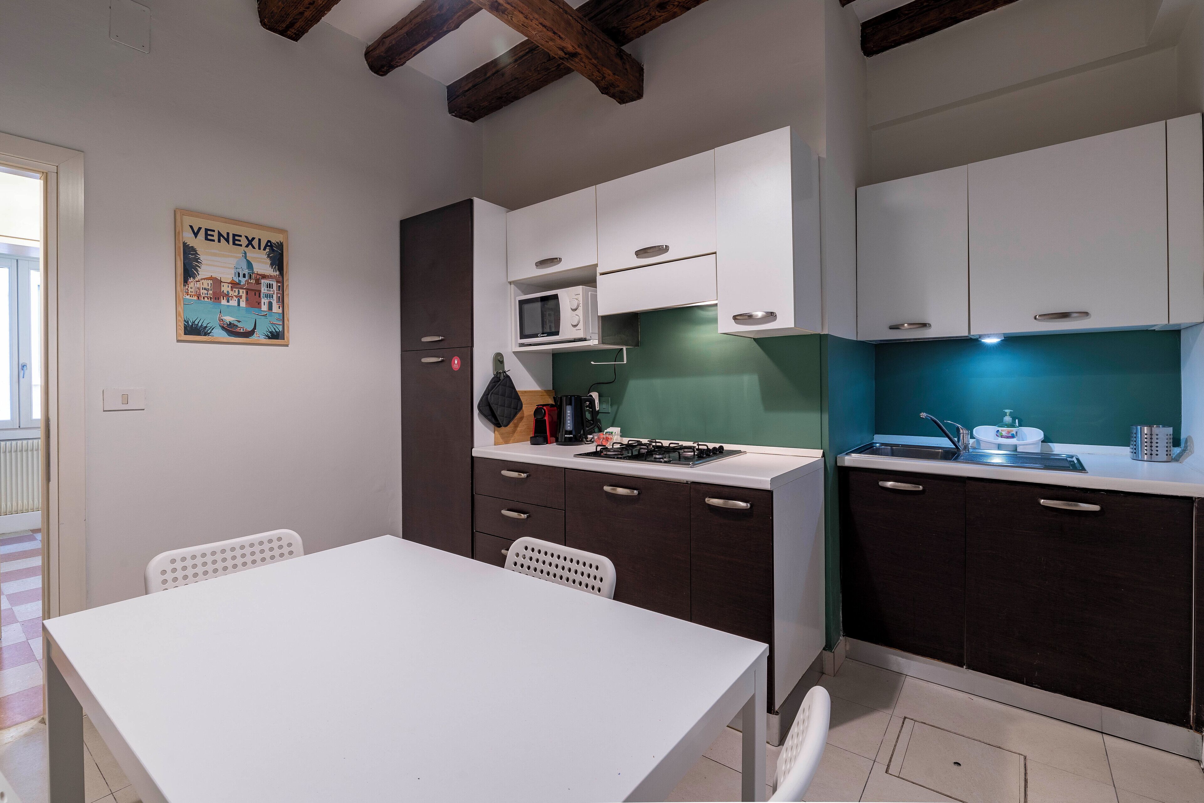 A compact kitchen with white and dark cabinets, a microwave, a gas stove, and a sink. There is a white table and two chairs. A poster of Venice is on the wall.