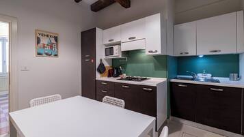 A compact kitchen with white and dark cabinets, a microwave, a gas stove, and a sink. There is a white table and two chairs. A poster of Venice is on the wall.