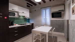 Cocina compacta con microondas, estufa y armarios blancos. Se observa una mesa de comedor con sillas y una ventana con cortinas.