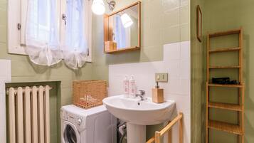 A bathroom with a washing machine, a sink, a mirror, and a wooden shelf.