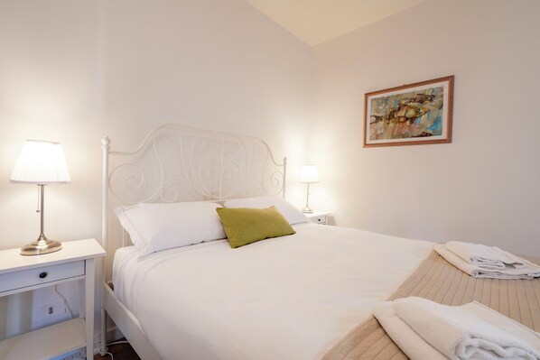 Una cama bien hecha con sábanas blancas y una almohada verde, dos lámparas de noche, una toalla doblada sobre la cama y una obra de arte enmarcada en la pared.