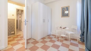 Una habitación con piso a cuadros, un armario blanco, dos sillas blancas, una mesita y un cuadro enmarcado en la pared.