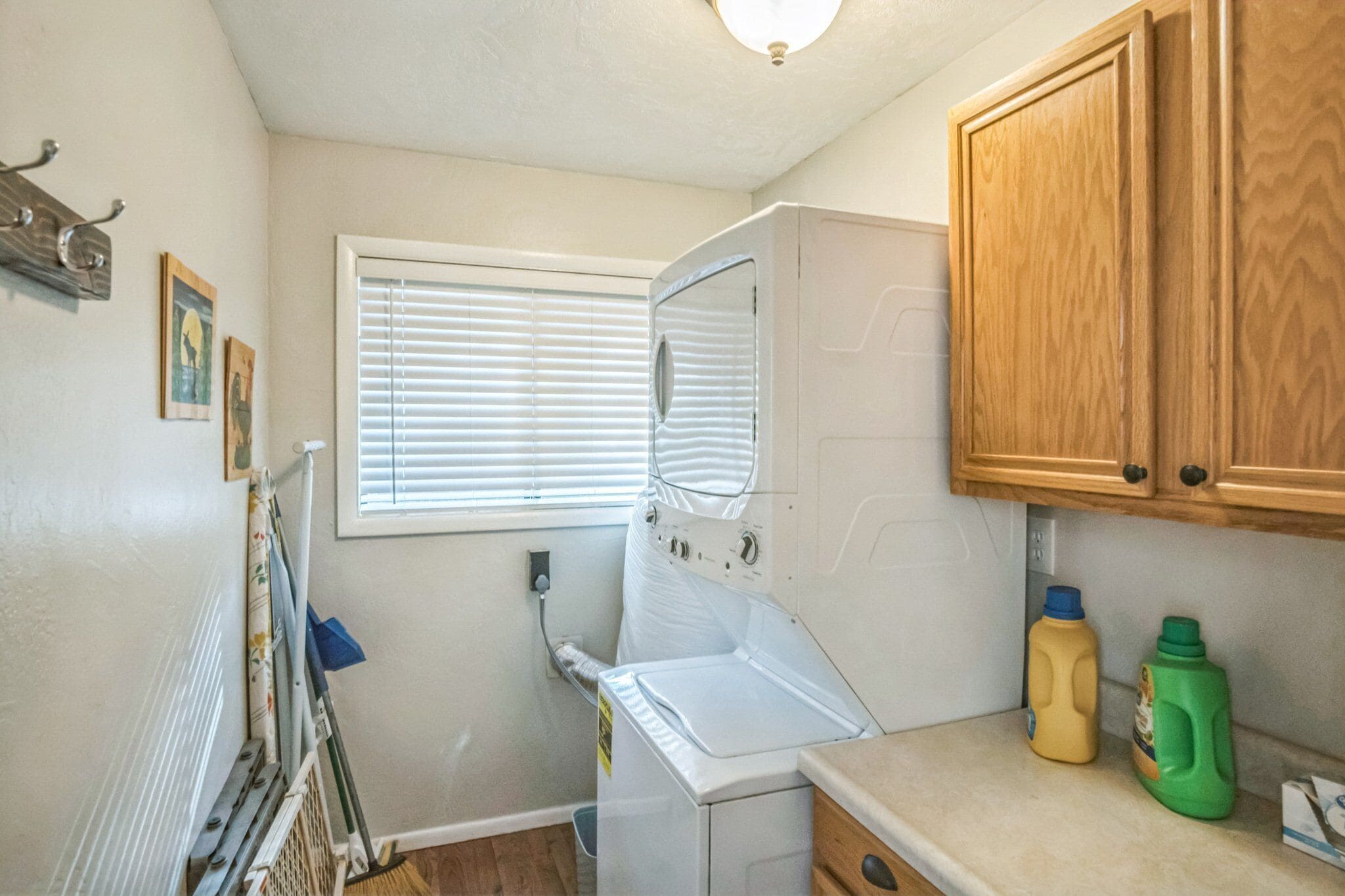 A laundry room with a dryer, a window with blinds, wooden cabinets, and two detergent bottles on the counter.