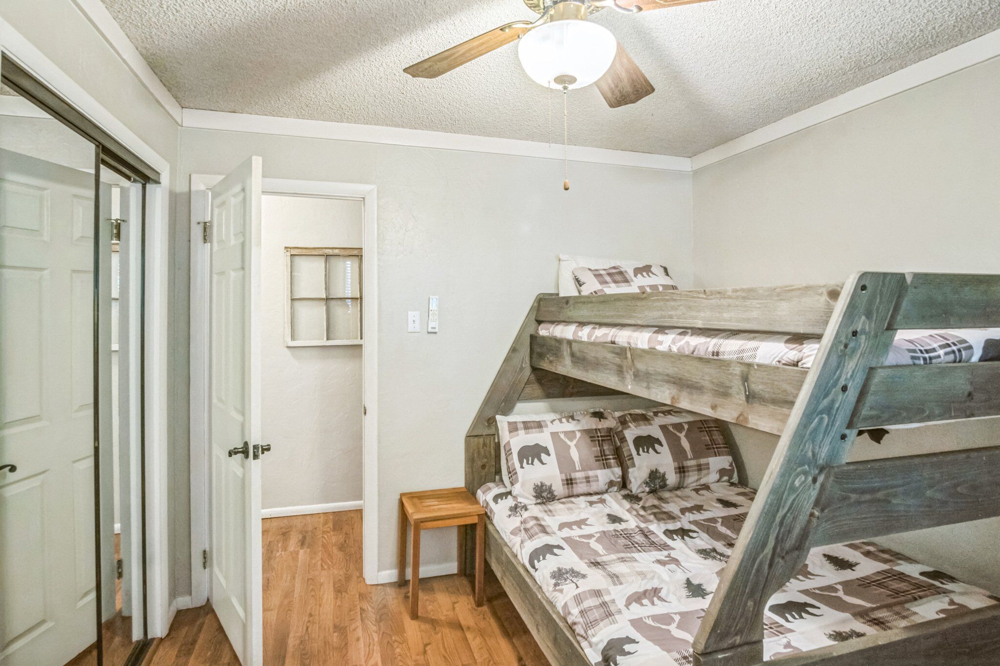 A room with a bunk bed, a ceiling fan, a small wooden stool, and a door leading to another room.