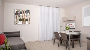 Ein modernes Wohnzimmer mit einem grauen Sofa, einem Esstisch mit Stühlen, einem weißen Vorhang und einem gerahmten Logo an der Wand.
