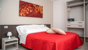 A modern bedroom with a red bedspread, a white headboard, a nightstand with a lamp, and a closet with hanging clothes. - CAMELOT APARTMENT RED3 (VERONA)