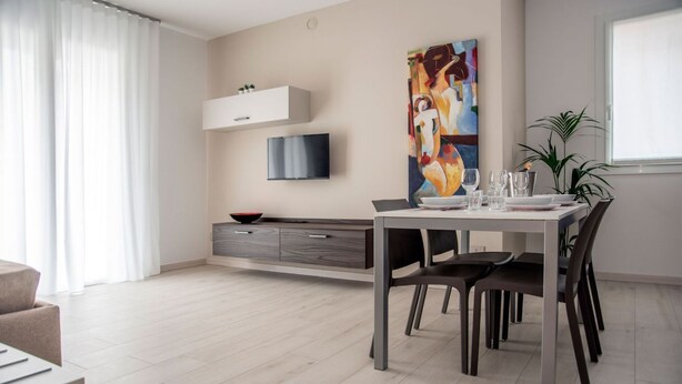 A modern living room with a dining table, chairs, a TV mounted on the wall, and a large abstract painting. - CAMELOT APARTMENT RED6 (VERONA)