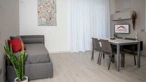 Ein modernes Wohnzimmer mit grauem Sofa, einem Esstisch mit Stühlen, einer Zimmerpflanze und einem gerahmten Kunstwerk an der Wand.