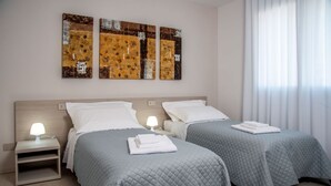Ein Hotelzimmer mit zwei Betten, jeweils mit einer grauen gesteppten Tagesdecke und weißen Kissen. An den Nachttischen stehen Lampen, und an der Wand über den Betten ist Kunst ausgestellt.