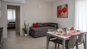 A modern living room with a grey sofa, a dining table set with plates, cups, and cutlery, and a potted plant. - CAMELOT APARTMENT RED4 (VERONA)