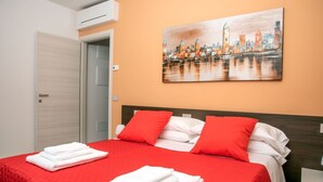 A hotel room with a bed, red and white bedding, a cityscape painting on the wall, and a door. - CAMELOT APARTMENT RED8 (VERONA)