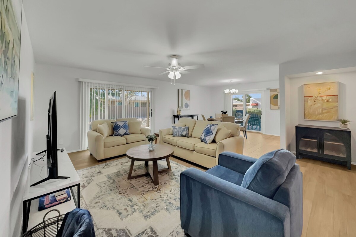 A modern living room with a beige sofa, a blue armchair, and a coffee table. There is a TV on a stand, a ceiling fan, and a painting on the wall.