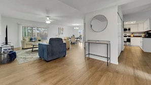 Een ruime woonkamer met een bank, fauteuil en salontafel, een eethoek met stoelen, en een keuken met witte kasten en apparatuur.