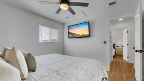 A bedroom with a bed, a ceiling fan, a TV on the wall, a window with blinds, and a hallway leading to another room. - Boulder Apache Junction (Apache Junction)