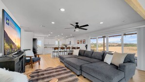 A spacious living room with a large gray sectional sofa, a wooden coffee table, and a fluffy rug. There is a kitchen area with white cabinets and a dining table. The room features a ceiling fan, recessed lighting, and large windows offering a view of the desert landscape. - Boulder Apache Junction (Apache Junction)