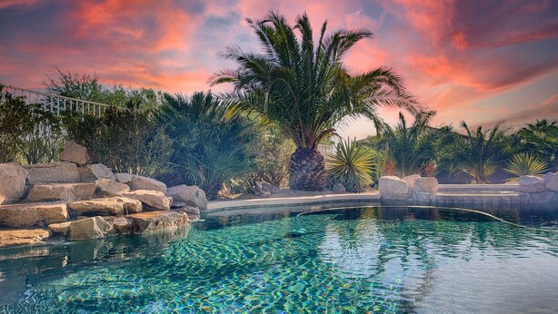 A clear swimming pool surrounded by rocks and palm trees, with a sunset sky in the background. - Cline Maricopa (Maricopa)