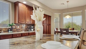 A kitchen with wooden cabinets, a granite countertop, a vase with dried flowers, and a dining area with a table and chairs. - Cline Maricopa (Maricopa)